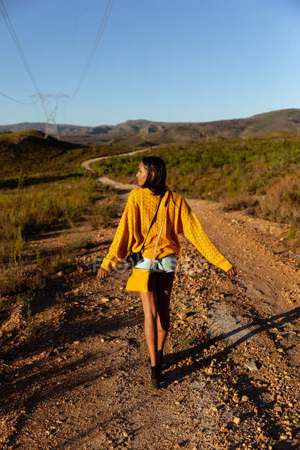 Visão traseira de uma jovem mulher de raça mista desfrutando de um passeio ao longo de uma trilha através de uma paisagem rural ensolarada em direção a montanhas no horizonte. Ela está usando shorts, com um top amarelo com uma bolsa e uma câmera . — Fotografia de Stock
