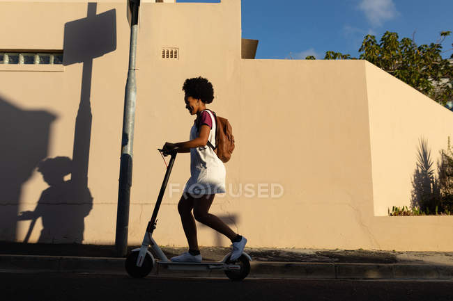 Vista laterale di una giovane donna mista che indossa uno zaino cavalcando uno scooter in una strada urbana al sole — Foto stock