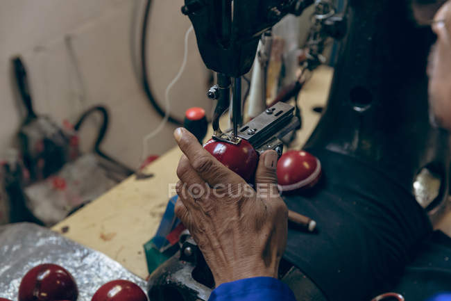 Fermer la main de l'homme travaillant sur la couture de la moitié extérieure en forme d'une balle de cricket à l'aide d'une machine à coudre dans un atelier d'une usine d'équipements sportifs . — Photo de stock