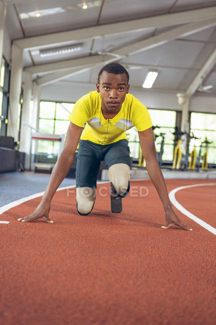 Vista frontal del atlético masculino afroamericano discapacitado en posición inicial en pista de atletismo en gimnasio - foto de stock