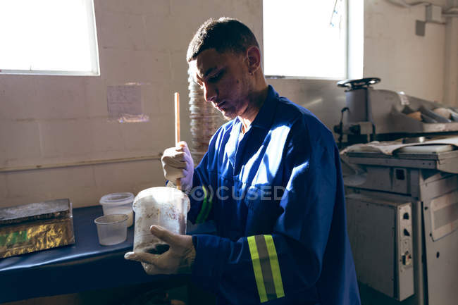 Side view close up of a young mixed race man wearing gloves and overalls preparing a mixture at a factory making cricket balls, with equipment visible in the background. — Stock Photo