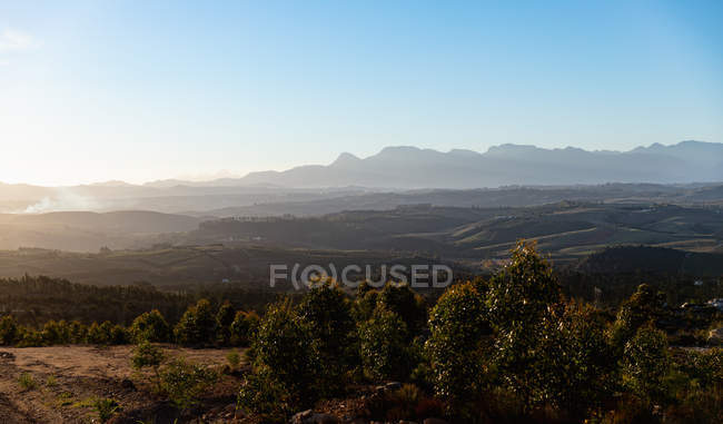 Wide view of an open natural landscape with trees in the foreground, distant mountains and sun setting on the horizon — Stock Photo