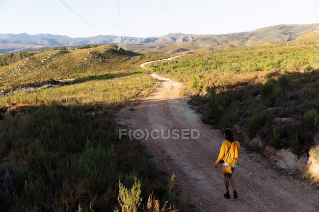 Visão traseira de uma jovem mista caminhando ao longo de uma trilha através de uma paisagem rural ensolarada em direção a montanhas no horizonte. Ela está usando shorts, com um top amarelo e uma bolsa . — Fotografia de Stock