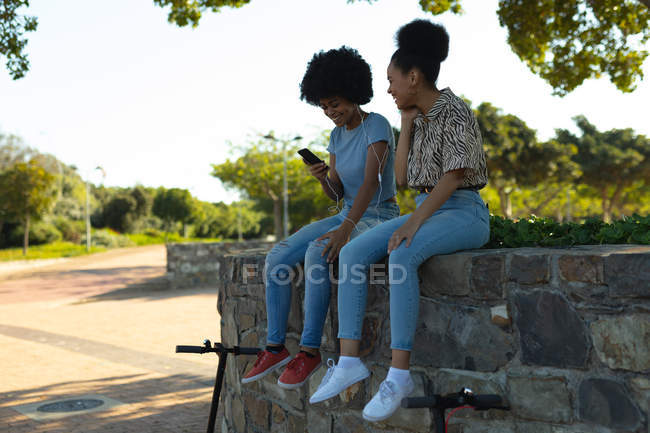 Side view of two smiling young adult mixed race sisters sitting on a wall in an urban park, using a smartphone and sharing earphones to listen to music, with their electric scooters parked below them — Stock Photo
