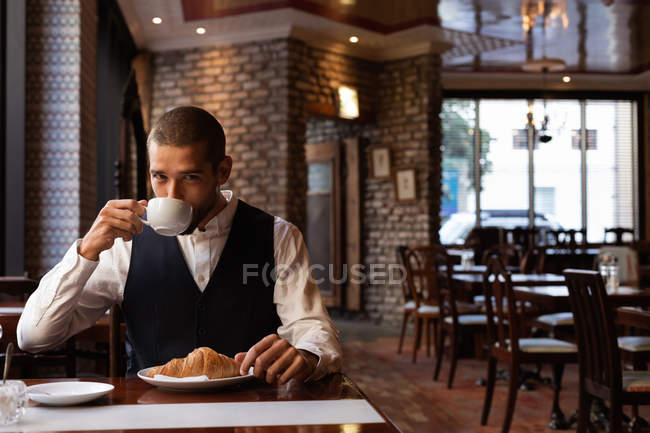 Vista frontal de cerca de un joven caucásico tomando café sentado en una mesa dentro de un café. Nómada digital en movimiento . - foto de stock
