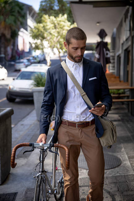 Vista frontal de cerca de un joven caucásico caminando, usando su teléfono inteligente y moviendo su bicicleta en una calle de la ciudad. Nómada digital en movimiento . - foto de stock