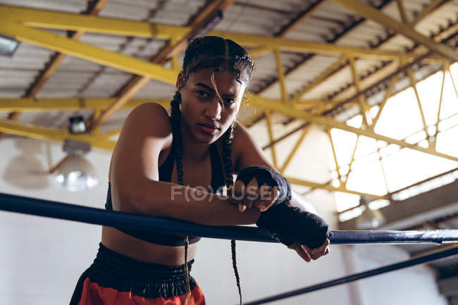 Visão de baixo ângulo do boxeador feminino apoiado em cordas e olhando para a câmera no ringue de boxe. Forte lutador feminino no treinamento de ginásio de boxe duro . — Fotografia de Stock