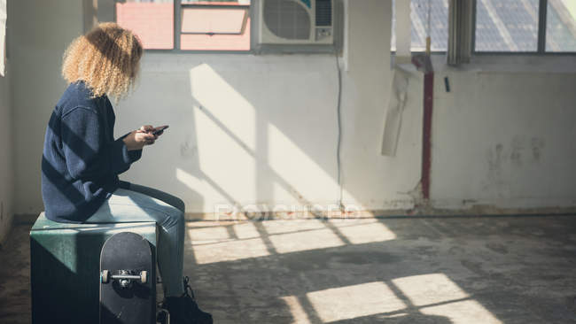Вид сбоку молодой белой женщины с вьющимися волосами с мобильного телефона рядом со скейтбордом, сидящей внутри пустого склада — стоковое фото