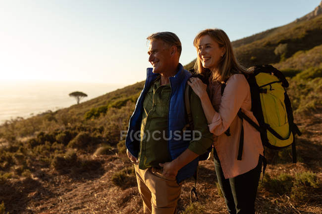 Seitliche Nahaufnahme eines reifen kaukasischen Mannes und einer Frau mit Rucksäcken, die anhalten, um die Landschaft bei Sonnenuntergang während einer Wanderung gemeinsam zu genießen. — Stockfoto