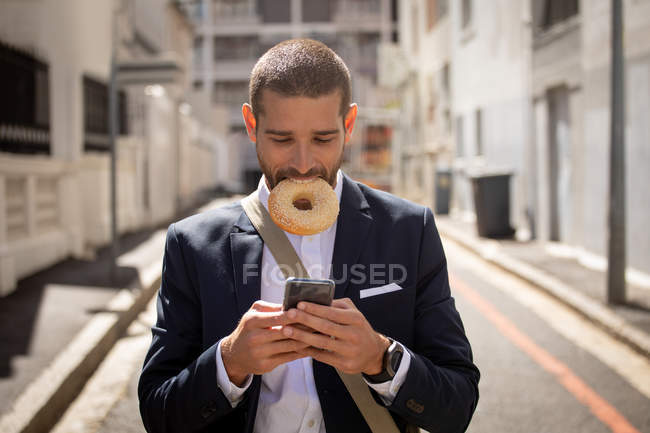 Вид спереду крупним планом молодого Кавказького чоловіка тримає кільцевий пончик в роті і використовуючи свій смартфон на міській вулиці. Цифрові кочівники на ходу. — стокове фото