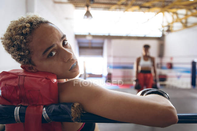 Close-up of female boxer looking at camera while resting in boxing ring at fitness center. Strong female fighter in boxing gym training hard. — Stock Photo