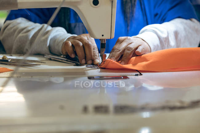 Закрыть руки женщины, шьющей швейной машинкой и оранжевой тканью на фабрике спортивной одежды . — стоковое фото