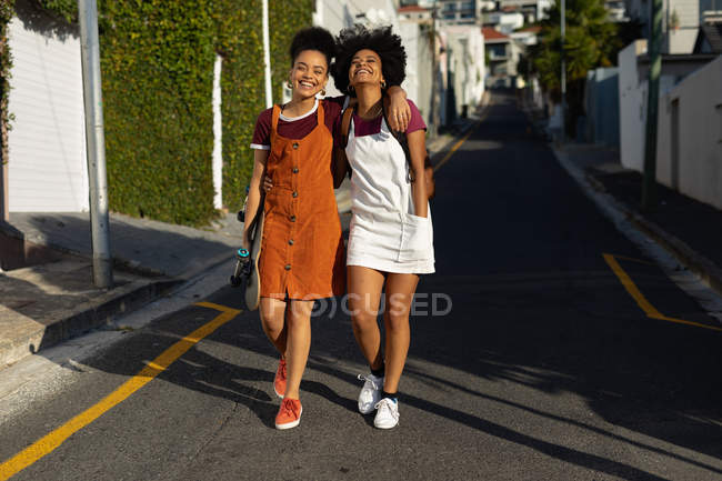 Vista frontal de dos hermanas adultas jóvenes de raza mixta con los brazos alrededor abrazándose mientras caminan en una calle bajo el sol, una con una mochila y la otra con un monopatín - foto de stock