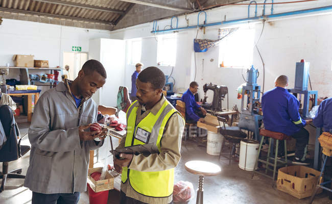 Front view of a young African American male manager holding a clipboard and talking with a young African American male worker in the workshop at a cricket ball factory. The worker is holding a cricket ball. — Stock Photo