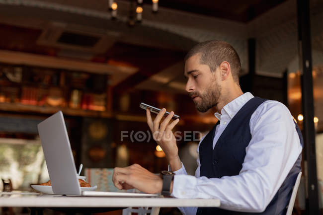 Vista lateral de perto de um jovem caucasiano fazendo um telefonema segurando seu smartphone na frente dele e usando um computador portátil, sentado em uma mesa dentro de um café. Digital Nomad em movimento . — Fotografia de Stock