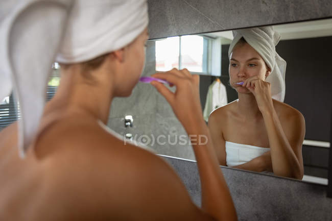 Über die Schulter einer jungen kaukasischen Frau, die sich die Zähne putzt, ein Badetuch trägt und ihr Haar in ein Handtuch gehüllt hat, das sich im Spiegel eines modernen Badezimmers spiegelt. — Stockfoto