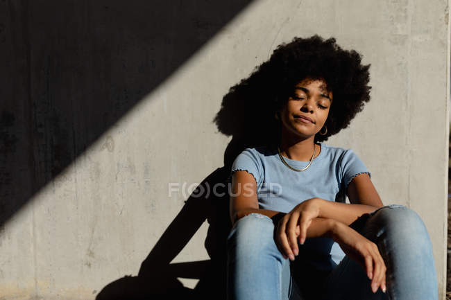 Портрет крупним планом молодої змішаної жінки, що сидить на стіні, розслабляючись з очима, закритими на відкритому повітрі на сонці — стокове фото