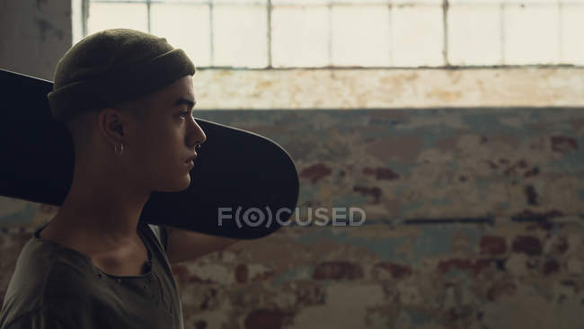 Вид сбоку на молодого латиноамериканца с пирсингом в тёмно-серой рубашке и шапочке, держащего скейтборд через плечо в пустом складе — стоковое фото