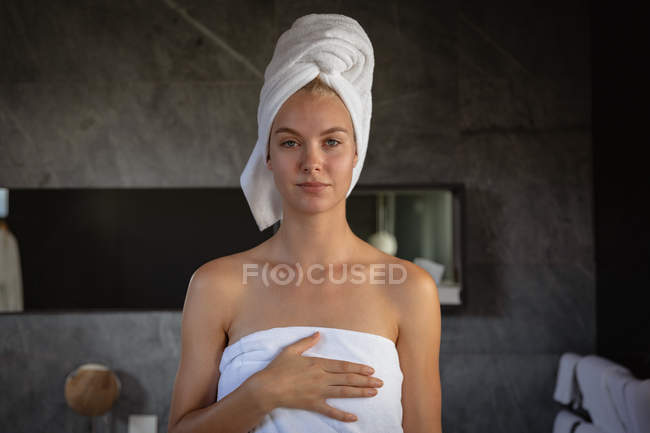 Porträt einer jungen kaukasischen Frau mit Badetuch und in ein Handtuch gehülltem Haar, die in einem modernen Badezimmer direkt in die Kamera blickt. — Stockfoto