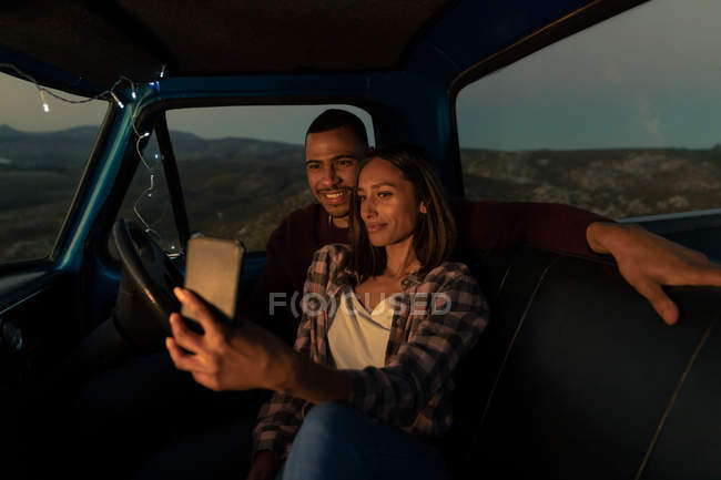 Vue rapprochée vers l'avant d'un jeune couple de course mixte assis dans leur voiture prenant un selfie au crépuscule lors d'un arrêt sur la route. L'intérieur de la voiture est éclairé avec des lumières à cordes . — Photo de stock