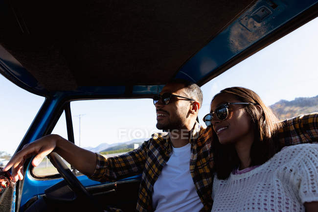 Vista a basso angolo di una giovane coppia mista seduta nel loro pick-up, sorridente e abbracciata durante un viaggio su strada. L'uomo sta guidando con il braccio intorno alla donna e entrambi indossano occhiali da sole e sorridono. — Foto stock