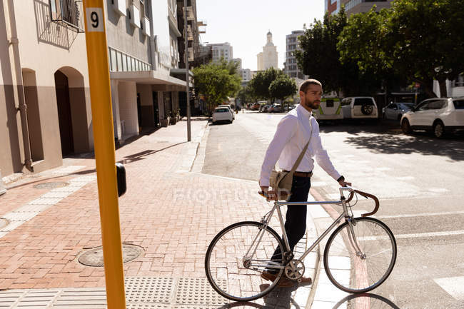 Vue latérale d'un jeune homme caucasien marchant avec son vélo traversant une rue de la ville. Nomade numérique en mouvement . — Photo de stock