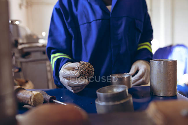 Вид спереди на середину человека в перчатках и комбинезоне, держащего ядро мяча и проверяющего его на заводе по производству мячей для крикета, в окружении оборудования . — стоковое фото