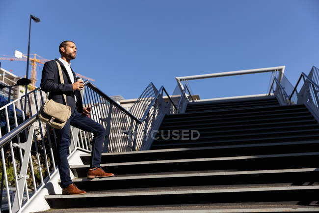 Vista lateral do jovem caucasiano carregando uma bolsa de ombro e segurando um café takeaway em pé em passos fora ao sol. Digital Nomad em movimento . — Fotografia de Stock