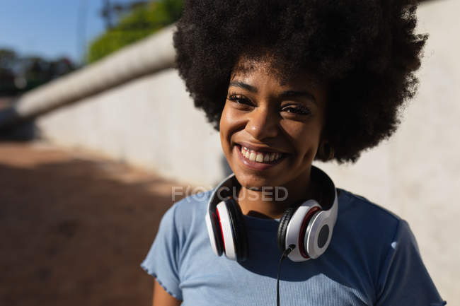 Portrait close up of a smiling young mixed race woman wearing headphones around her neck in a sunny urban park — Stock Photo