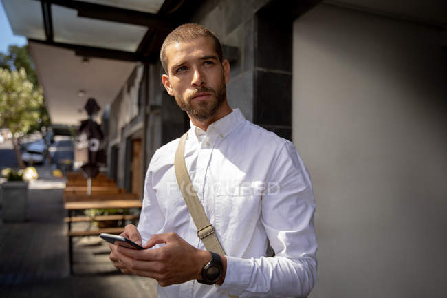 Vista frontal de perto de um jovem caucasiano usando um smartphone, de pé no pavimento em uma rua da cidade e olhando para longe. Digital Nomad em movimento . — Fotografia de Stock