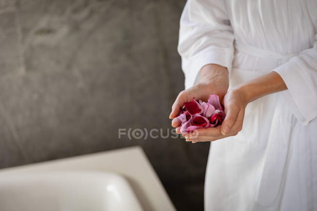 Середина жінки в халаті, що тримає кілька пелюсток троянд, стоїть поруч з ванною в сучасному туалеті . — стокове фото