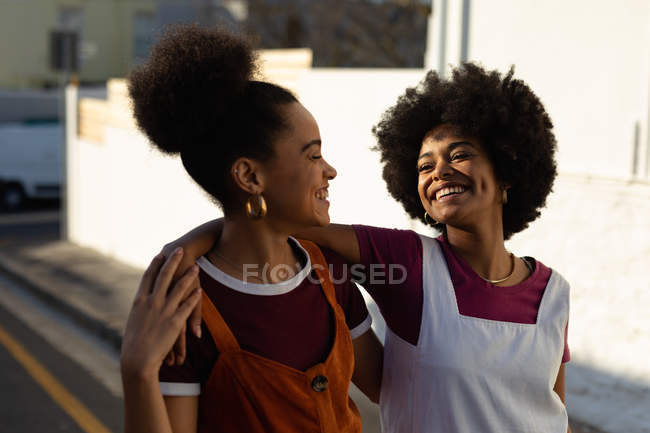 Вид спереди на двух взрослых сестер смешанной расы, которые улыбаются и смотрят друг на друга, идя по улице на солнце — стоковое фото