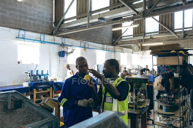Frontansicht zweier junger afrikanisch-amerikanischer männlicher Kollegen im Gespräch neben einer Maschine in einer Cricketballfabrik, einer hält einen gelben Ball, im Hintergrund arbeiten Menschen an Maschinen. — Stockfoto