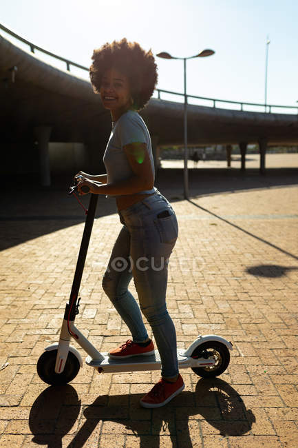 Vista lateral de perto de uma jovem mulher de raça mista montando uma scooter elétrica em um parque urbano, olhando para a câmera sorrindo, retroiluminado com brilho da lente — Fotografia de Stock