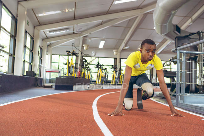 Vue de face de l'athlète masculin afro-américain handicapé en position de départ sur piste de course dans le centre de remise en forme — Photo de stock