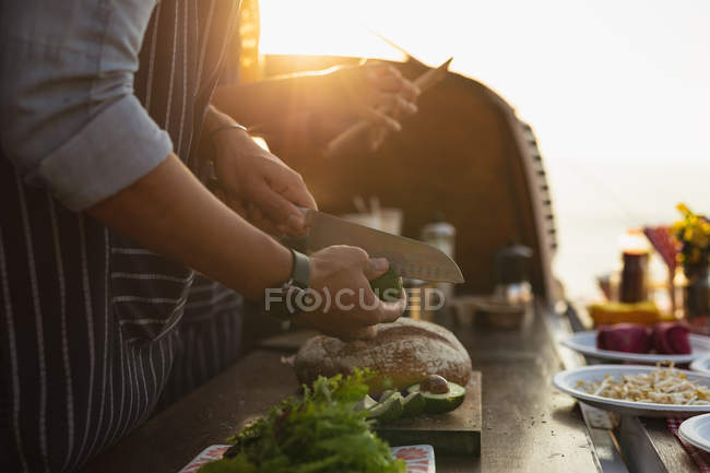 Side view mid section of couple busy preparing food standing in an open top van offering a range of takeaway food for sale, the woman is holding a tablet computer, they are backlit by sunlight — Stock Photo