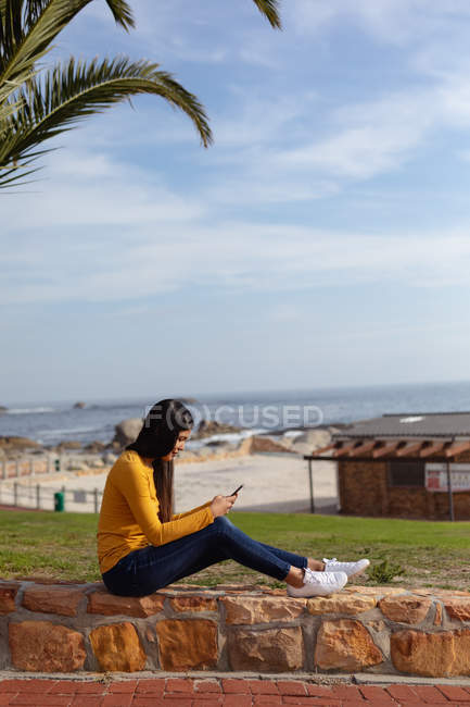 Вид збоку на молоду змішану жінку, яка сидить на вулиці на стіні, використовуючи смартфон, пальму і вид на море на задньому плані — стокове фото