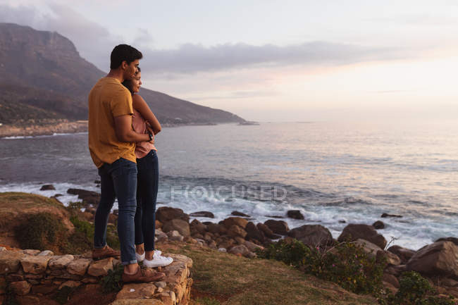 Vista laterale di una giovane coppia mista in piedi su una spiaggia che abbraccia e guarda verso il mare al tramonto — Foto stock