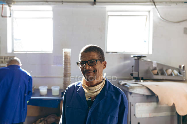 Retrato de un hombre de raza mixta de mediana edad con gafas, mirando a la cámara y sonriendo a una fábrica haciendo pelotas de cricket, con un compañero de trabajo trabajando en el fondo . - foto de stock