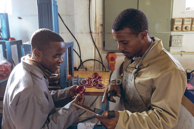 Vista laterale elevata di un giovane manager afroamericano con in mano un tablet e che parla con un giovane lavoratore afroamericano in officina in una fabbrica di palline da cricket . — Foto stock