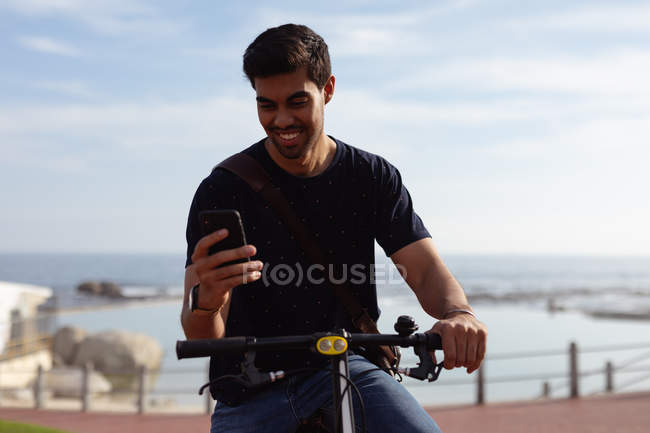 Вид спереди на молодого смешанного гонщика, сидящего на велосипеде с помощью смартфона в солнечный день, вид на море на заднем плане — стоковое фото