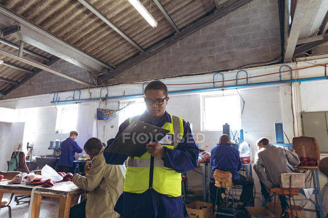 Front view of a young mixed race man wearing glasses writing on a clipboard at a cricket ball factory, while colleagues work on the production line at workbenches in the background. — Stock Photo