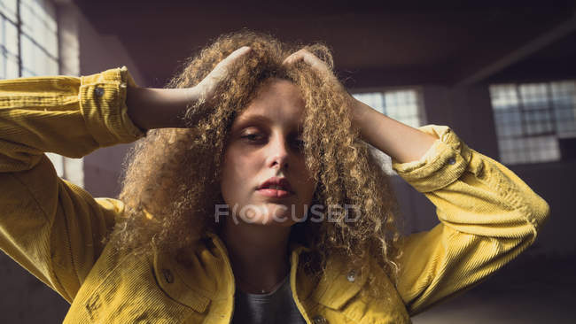 Front view of a young Caucasian woman with hands on curly hair wearing a yellow jacket over a grey shirt looking intently at the camera inside an empty warehouse — Stock Photo