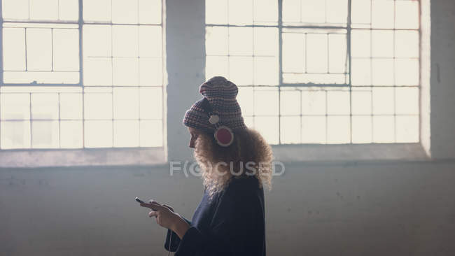 Вид сбоку на молодую белую женщину с вьющимися волосами в черных длинных рукавах и наушниках поверх шапки во время использования мобильного телефона внутри пустого склада — стоковое фото