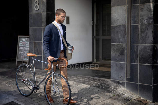 Vista lateral de un joven caucásico caminando sosteniendo una taza de comida para llevar y moviendo su bicicleta en una calle de la ciudad. Nómada digital en movimiento . - foto de stock