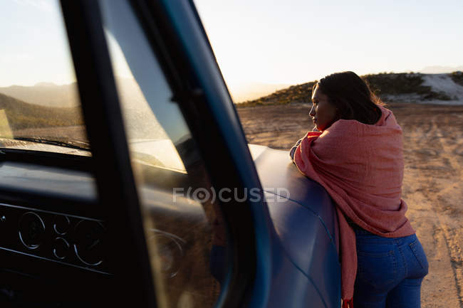 Vista lateral de una joven mujer de raza mixta apoyada en el capó de una camioneta y disfrutando de la vista al atardecer durante una parada en un viaje por carretera - foto de stock
