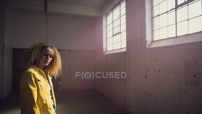 Vista lateral de uma jovem caucasiana com cabelo encaracolado vestindo uma jaqueta amarela sobre uma camisa cinza olhando atentamente para a câmera dentro de um armazém vazio — Fotografia de Stock