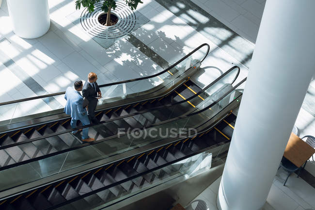 Vue grand angle des hommes d'affaires interagissant les uns avec les autres tout en descendant sur l'escalier roulant dans le bureau
. — Photo de stock