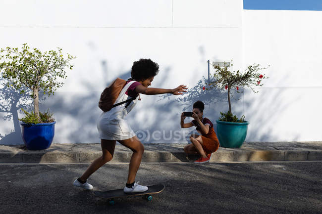 Вид сбоку на молодую женщину смешанной расы, катающуюся на скейтборде на городской улице, в то время как на заднем плане ее сестра-близнец опускается на колени, используя смартфон, чтобы сфотографировать — стоковое фото