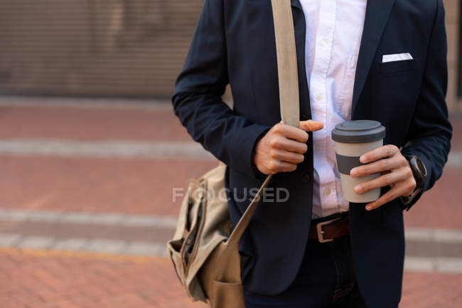 Front view mid section of man wearing a jacket, carrying a shoulder bag and holding a takeaway coffee, standing on a city street. Digital Nomad on the go. — Stock Photo